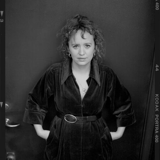 Sort-hvidt portræt af forfatteren Olga Ravn i mørk kjole med bælte, stående foran en mørk baggrund.