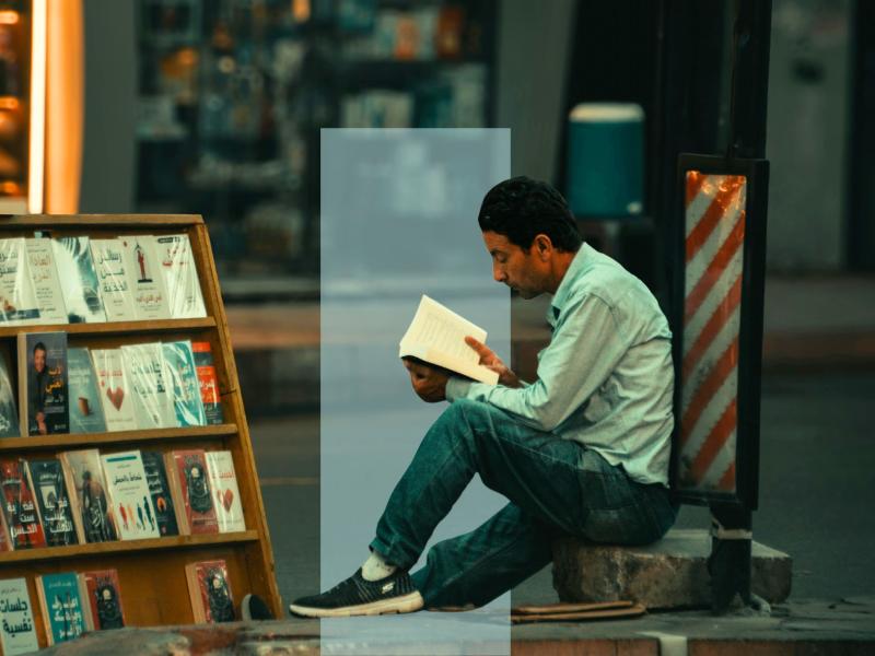 En mand sidder på en kantsten og læser i en bog ved siden af et bogstativ.